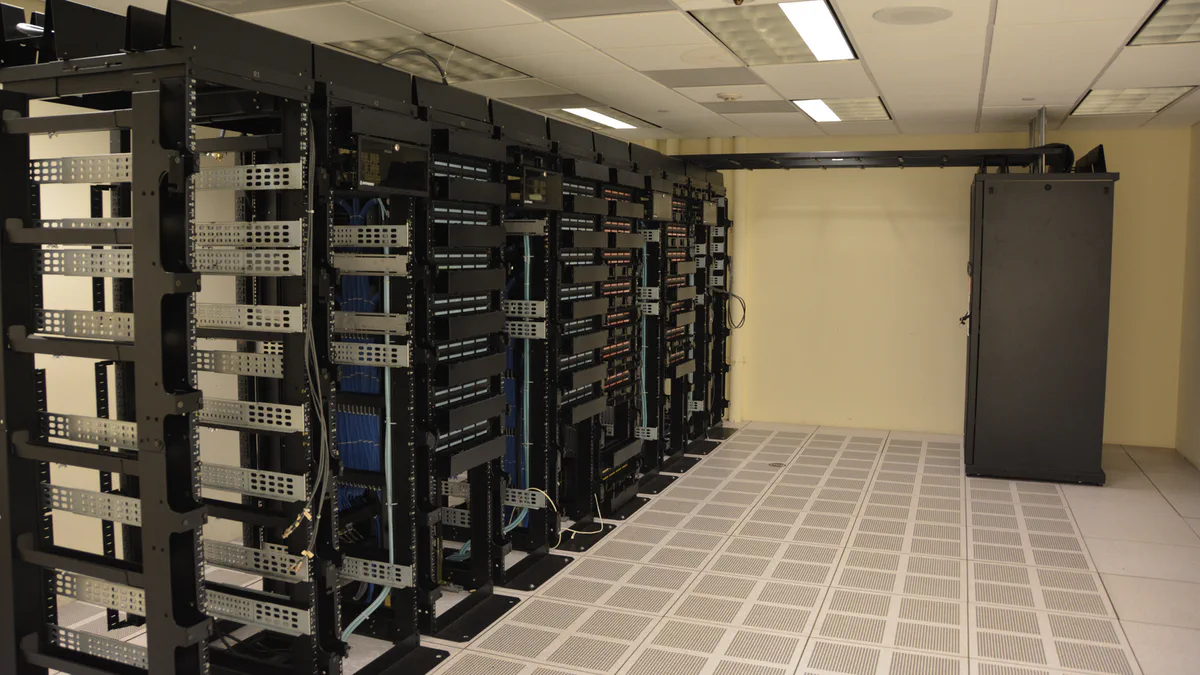 A corridor of server racks in a data center, with rows of dark equipment cabinets and cable runs under bright overhead lighting.