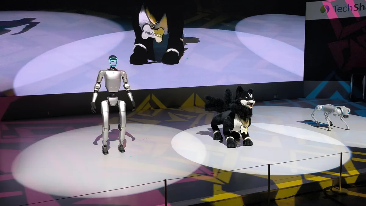 Three robots on a stage at the Japan Mobility Show 2025: a Unitree G1 humanoid, a small fox-like robot, and a Unitree Go2 four-legged robot.