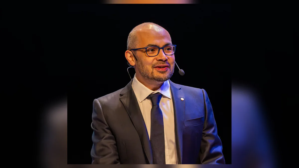 Portrait photograph of Demis Hassabis in a dark blazer and open-necked shirt against a plain background.