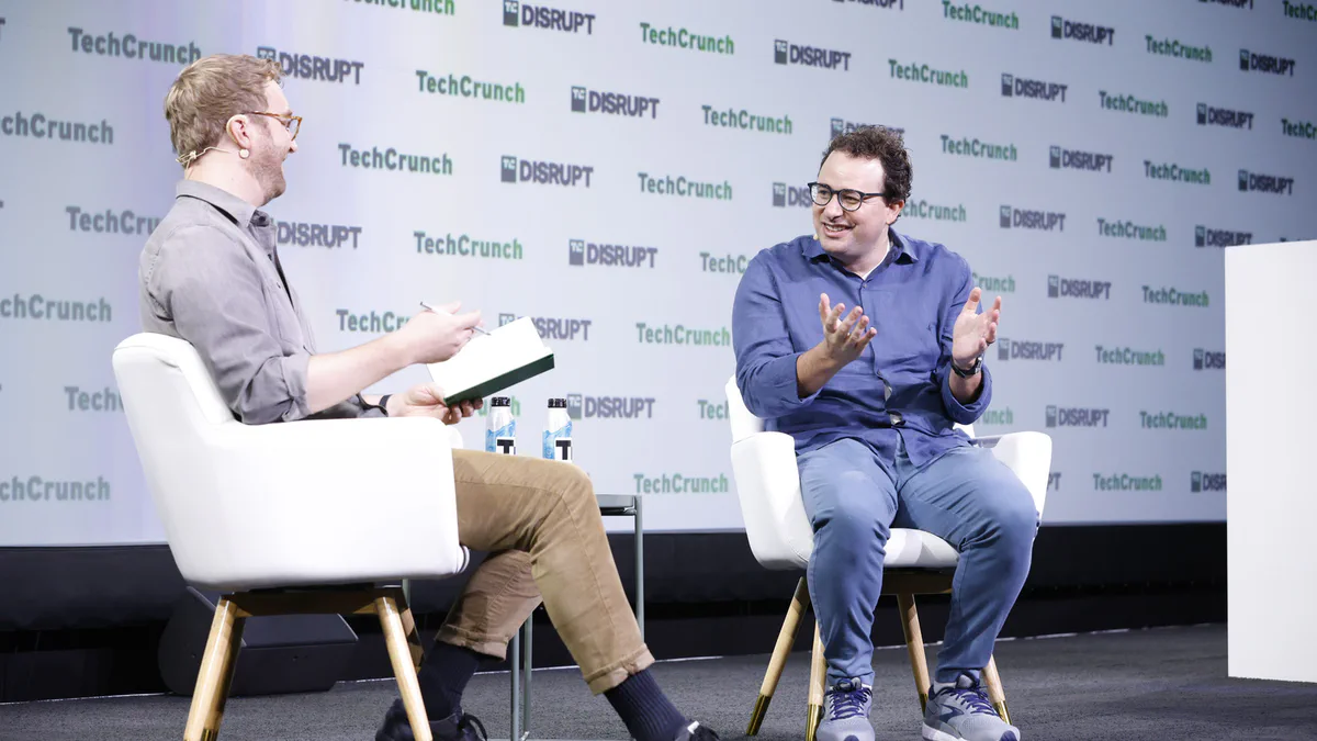 Dario Amodei seated in a white armchair on the TechCrunch Disrupt stage, gesturing while speaking with an interviewer.