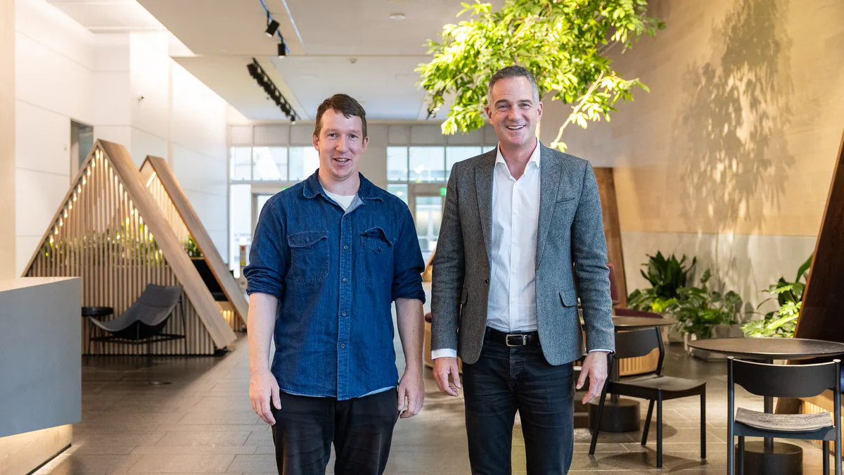 Two men stand in a bright office lobby with slatted wooden seating alcoves, a large indoor tree and dark chairs.