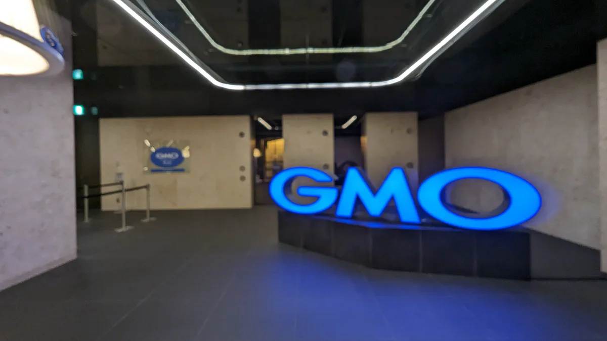 An illuminated blue GMO sign stands on a dark plinth in a lobby with pale stone walls, a metal barrier and a polished tiled floor.