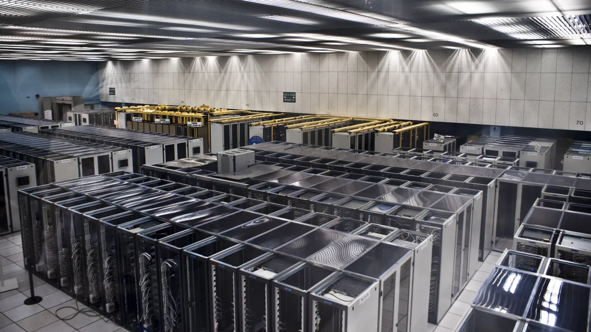 Rows of gray server cabinets with glass doors fill a brightly lit data hall, yellow cable trays running above one row and perforated metal ceiling panels overhead.